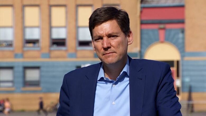 A man with brown hair in a suit stands in front of a school.