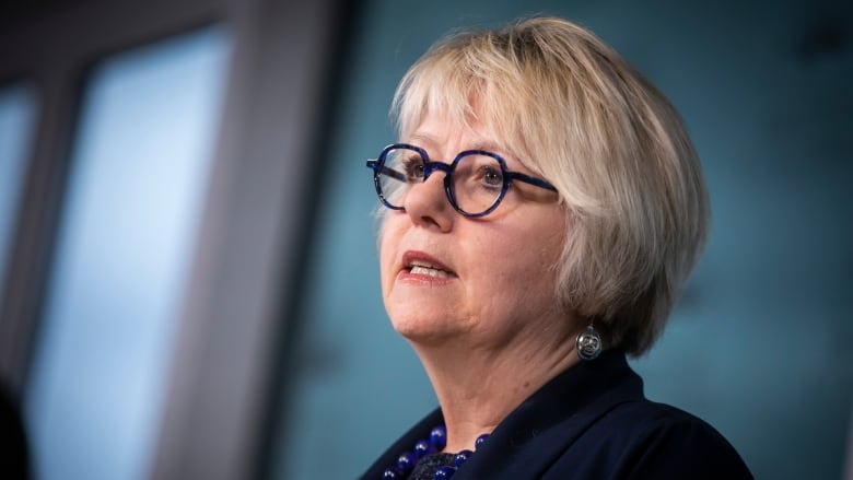 A white woman with short white hair, and blue glasses and a blue beaded necklace, speaks at a news conference.