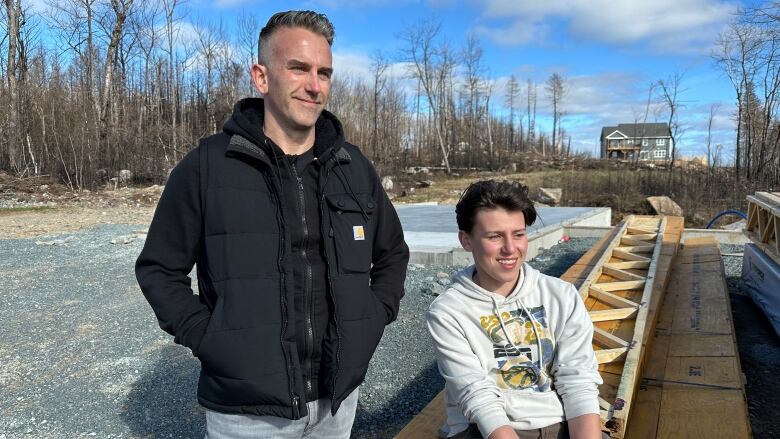 A man wearing a black jacket is standing by his son who is wearing a white hoodie and sitting on construction materials on their Upper Tantallon home.