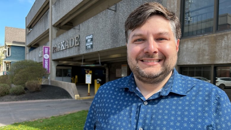 A man with short brown hair and a blue dotted shirt standing in front of a parking structure.