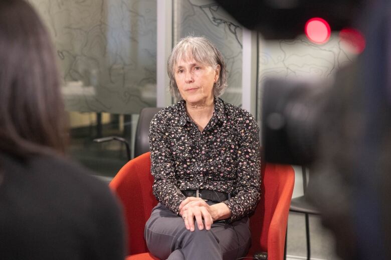 A woman sits in a red chair and faces a video camera.