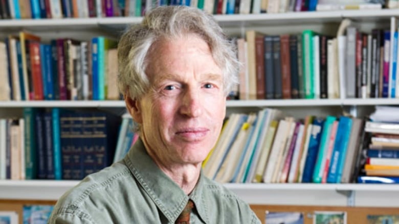 A man with white hair is looking into the camera. He is sitting in front of a shelf full of books.