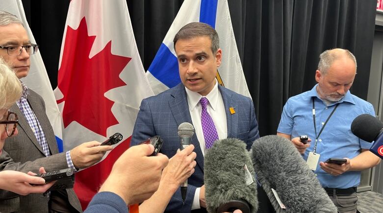 A man in a suit and tie stands in front of microphones while people stand to either side of him.