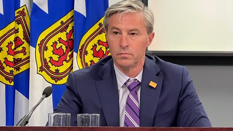 A man wearing a suit and tie sits at a desk with a microphone in front of Nova Scotia flags.