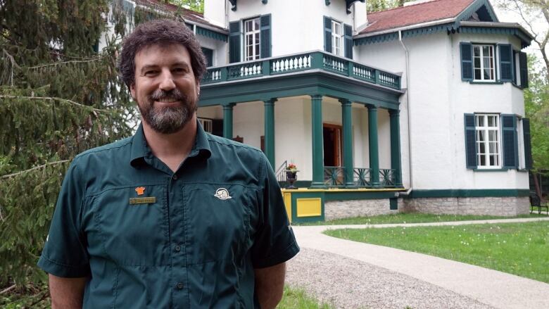 A man with bushy brown hair and a beard stands in front of a fancy white house with green trim.