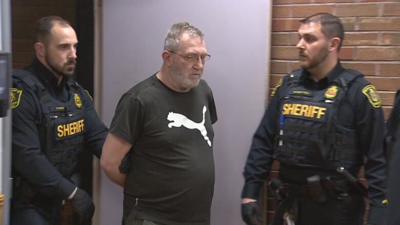 A man with a black shirt is flanked by two sheriff's officers.