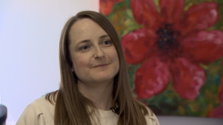 A woman with straight, brown hair stands in front of a red floral mural.