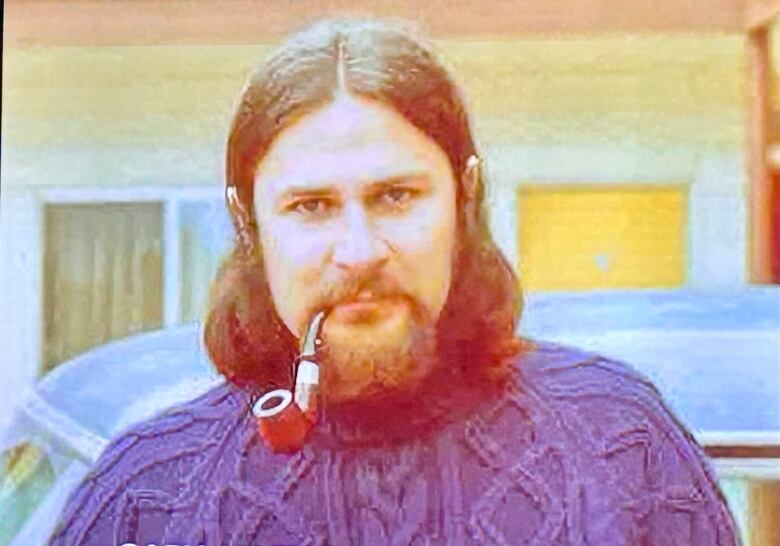 Old photograph of long-haired, bearded man with a purple sweater and a pipe.