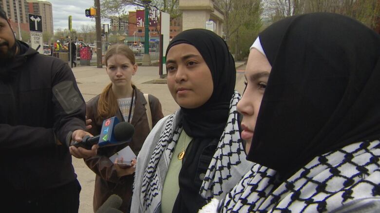 Two women with hijabs are standing on the sidewalk outside, in front of several microphones held by reporters.