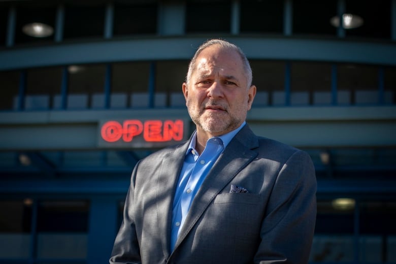 A white man wearing a grey sports jacket and a blue shirt stands in front of a red 'open' sign.