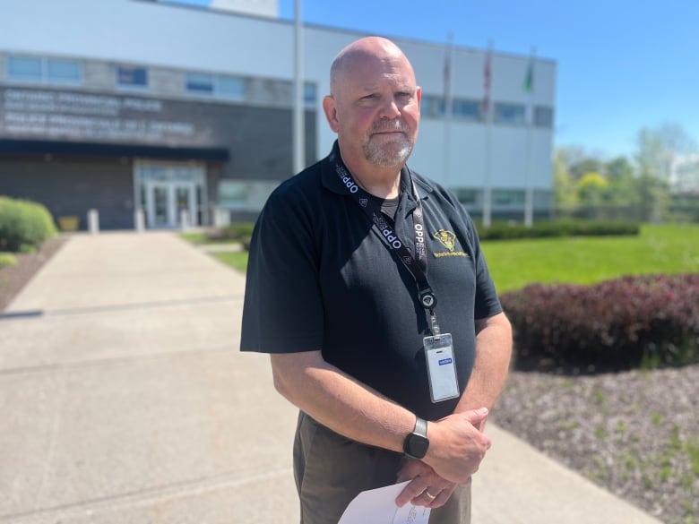 A man in a black OPP shirt stands in front of a building and frowns.