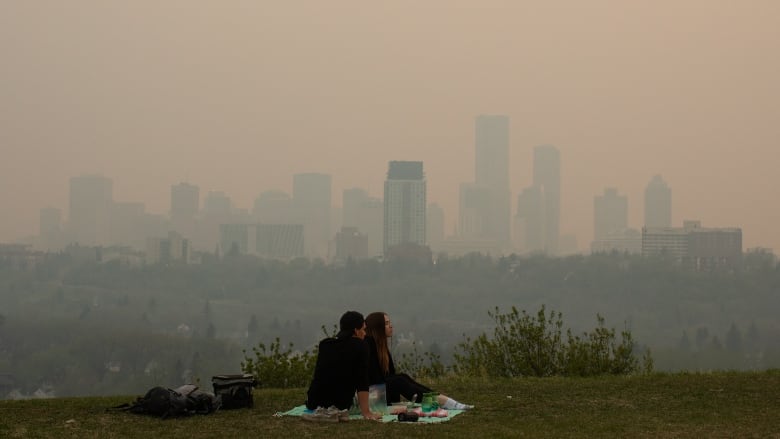 A couple sits having a picnic in front of a smoky downtown skyline.