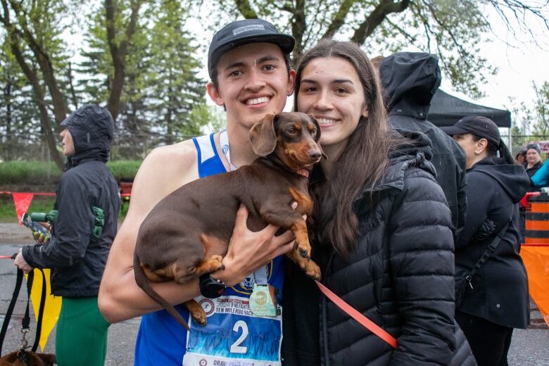 Two people are seen standing together at the end of a race, holding a dachshund.