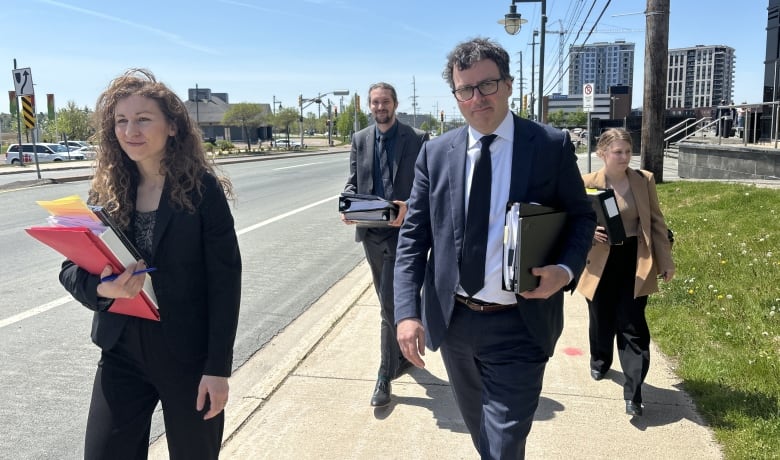 A woman with curly hair holding a binder and a man with files walk ahead of two other people on a sidewalk.