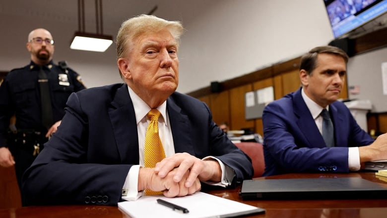 A man in a blue suit with an orange tie sits at a desk with his hands folded.