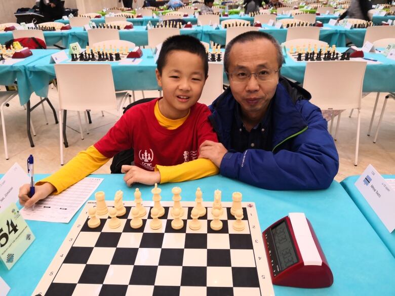 A man and a young boy pose in front of a chess board.