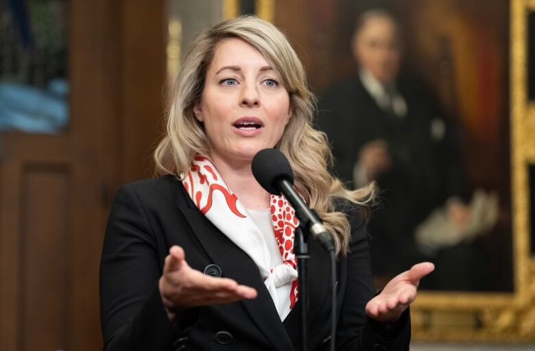 A woman in a black jacket and white and red scarf speaks into a microphone.