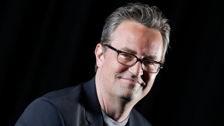 A man wearing glasses and a suit jacket smiles in front of a black background. 