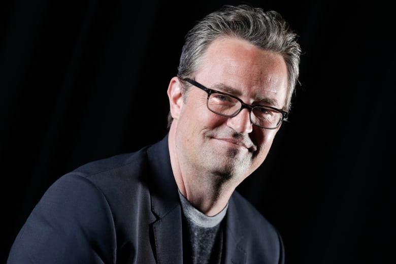 A man wearing glasses and a suit jacket smiles in front of a black background. 