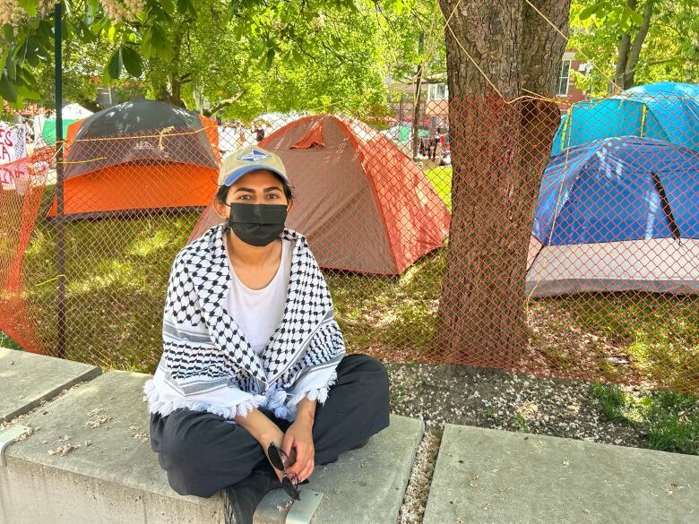 A woman sits on a concrete curb wearing a mask. 