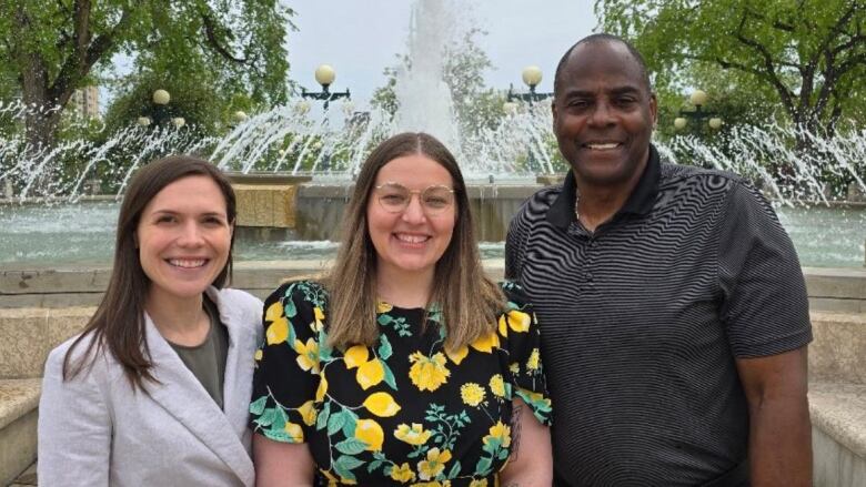 A woman in a grey blazer, a woman in a flowery yellow and green dress and a man in a black shirt stand side-by-side, in front of a large outdoor fountain.