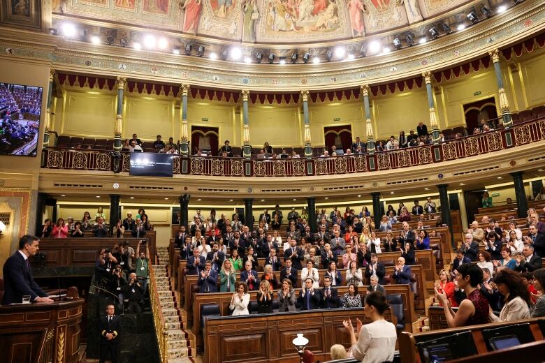 A man at a podium speaks at a podium in front of a crowded legislature.