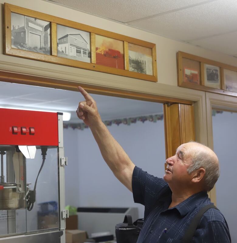 An older man points at old black and white photos on the wall.