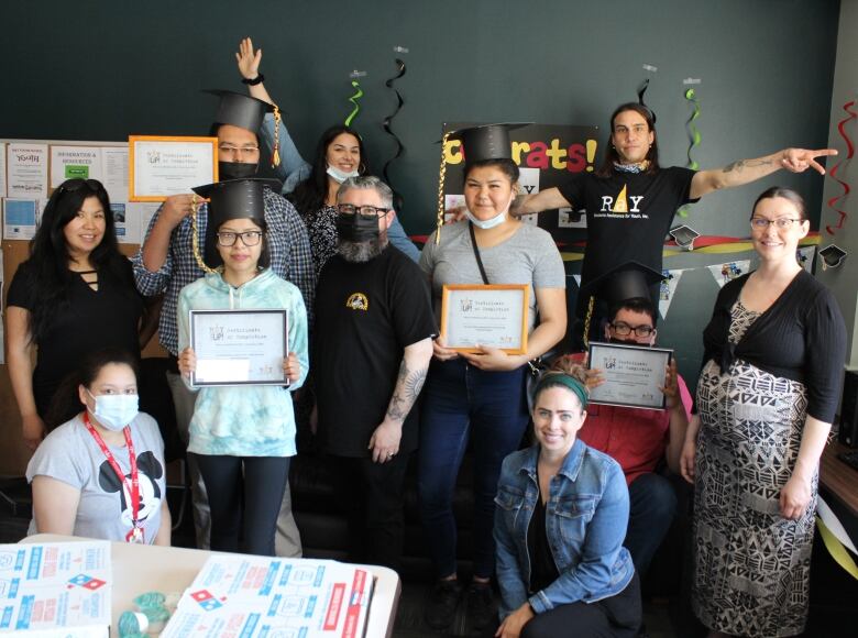 Students stand in a classroom holding framed certificates. Some students wear a graduation cap. 