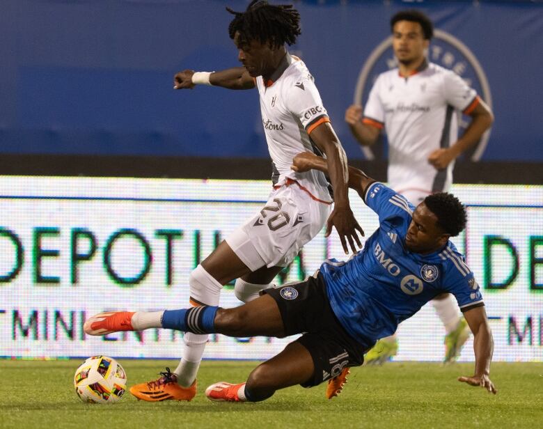 A soccer player wearing white has the ball while a player wearing blue slides in.