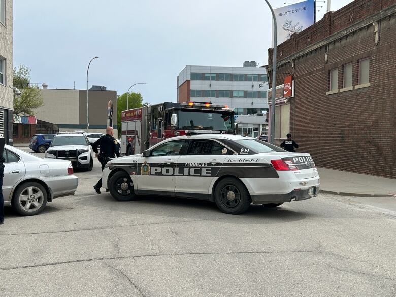 Two police officers stand near a police vehicle and firetruck on the street. 