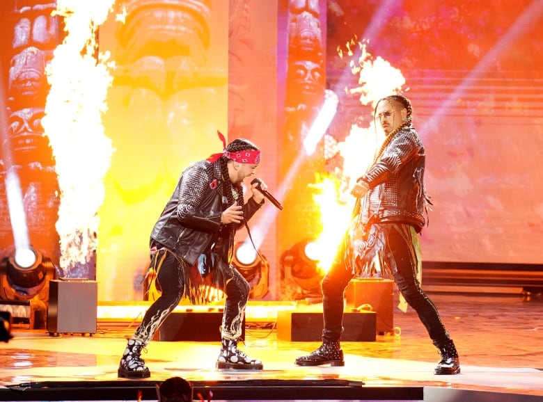 Two men perform on stage with flames in the background.