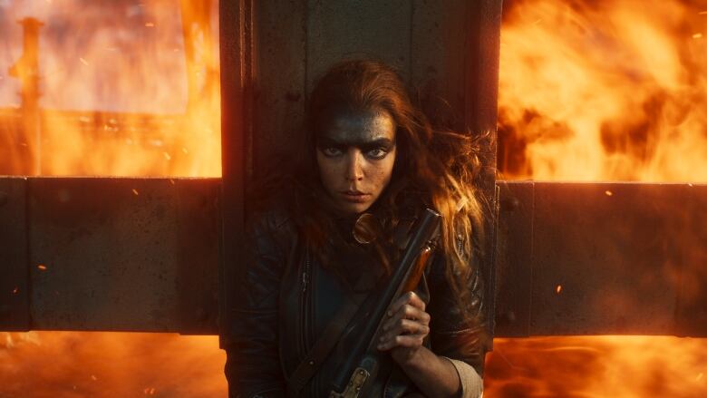 A woman with black makeup on her forehead braces herself in front of an iron grate. She is holding a shotgun. Behind the grate is a wall of fire. 