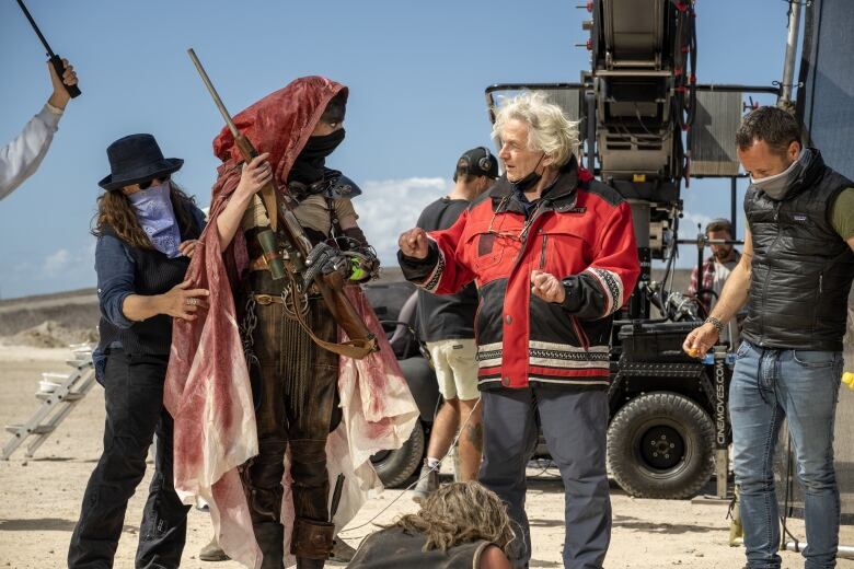 A woman wearing robes and dirty brown clothes speaks to a man wearing a red jacket. Behind them is a camera crew and a large vehicle mounted video camera. 