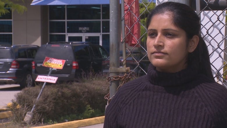 A South Asian woman in a black sweater stands in front of a chain link fence in a parking lot. She is framed in the image as though she's doing a TV interview.