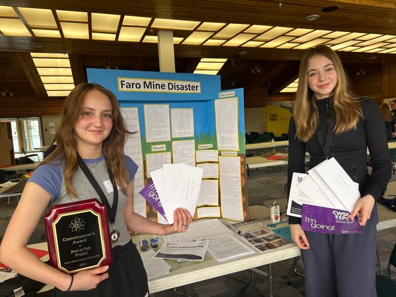 Two girls holding envelopes and a plaque stand in front of a poster board stand labelled, Faro Mine Disaster.