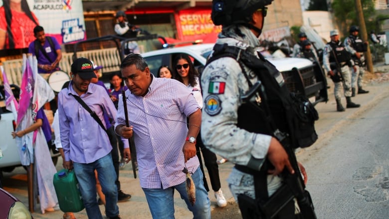 A man in a pink shirt walks past a member of the national guard holding a gun.