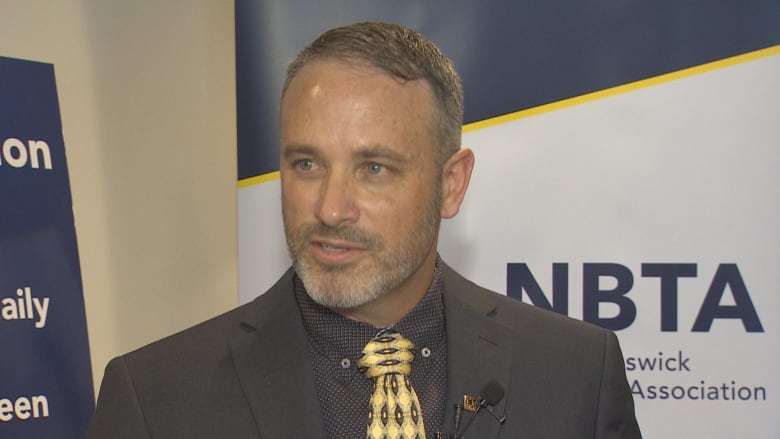 A person wearing a tie and blazer stands in front of a white, yellow and blue banner with the letters 