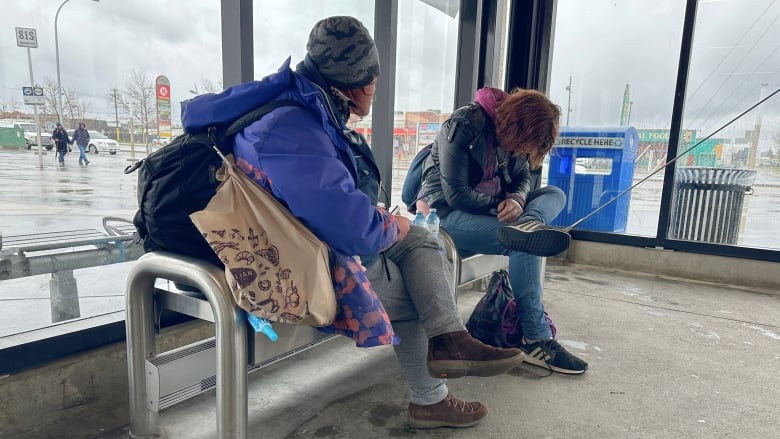 Two people sit on a bench. The woman closest to the camera has a large backpack and many bags.