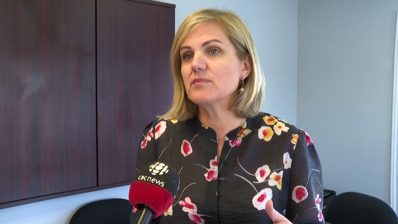 A woman with shoulder-length blonde hair and a black and floral shirt speaks into a microphone in an office setting.