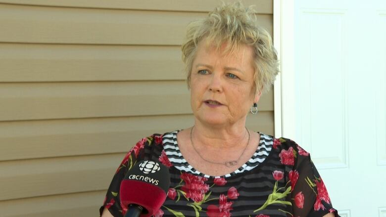 A woman with short blonde hair and a black and floral patterned shirt speaks into a microphone outside of a yellow coloured building.