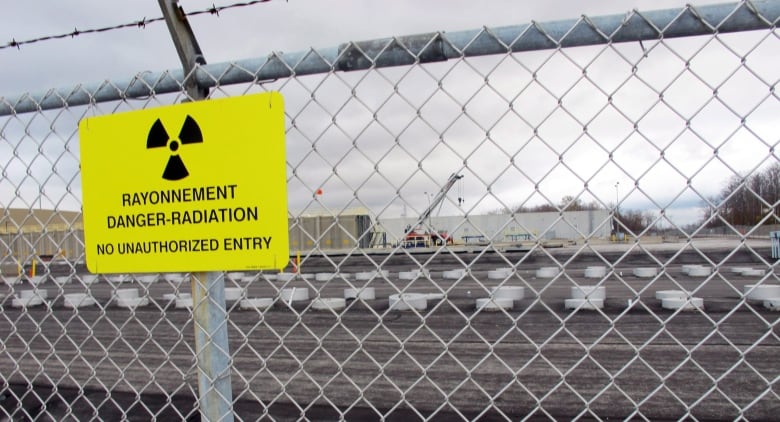 A sign indicating a radiation danger is posted on a chain-link fence, topped with barbed wire. A complex of buildings is visible in the background.