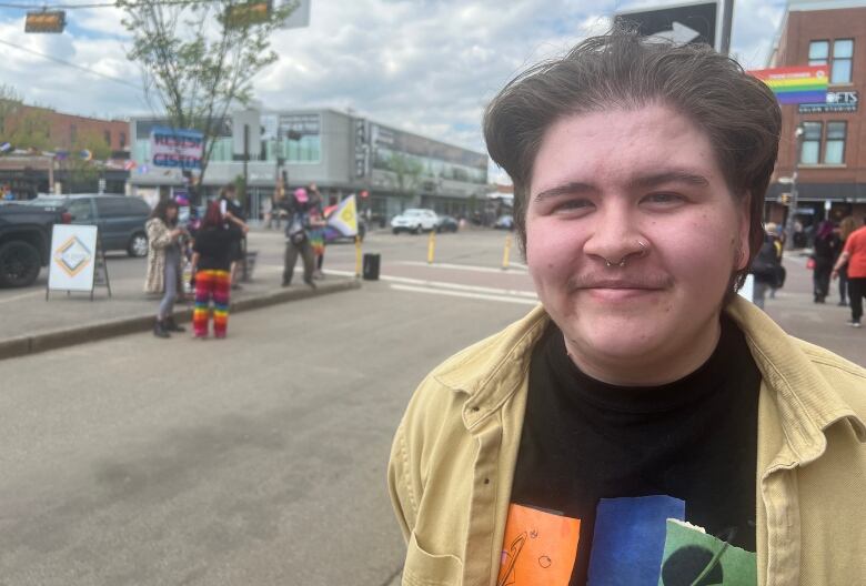 A person stands on a busy Edmonton street corner. They have a nose piercing and they're wearing button up shirt over a T-shirt. In the background, members of the queer community dance on Whyte Avenue in Edmonton.