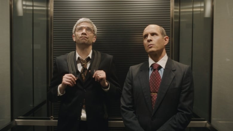 Still from the CBC limited series BlackBerry. Actors Jay Baruchel and Glenn Howerton appear standing in a medium shot, facing the viewer. They are two white men wearing dark suits. They are standing side by side in an elevator looking upward.