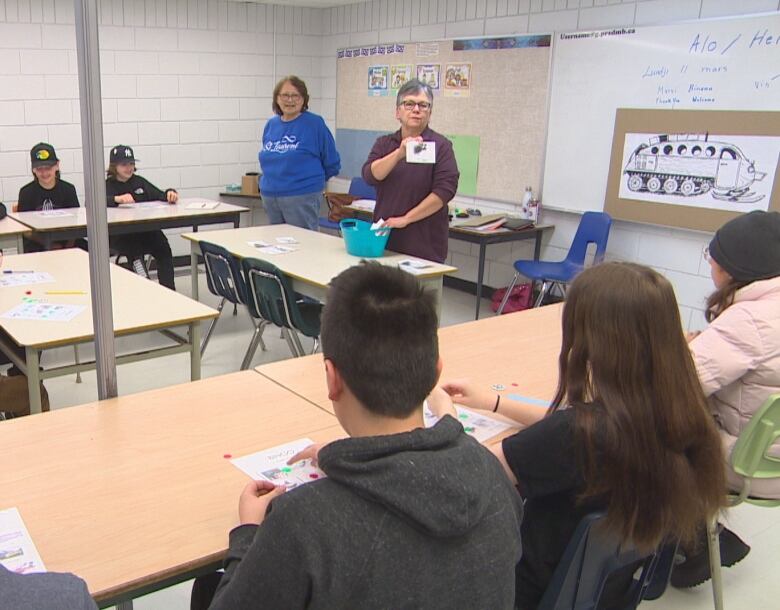 Two women stand in front of a classroom, one of them holding a flashcard. 