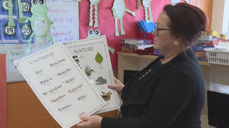 Kim Colwill hold up two posters in Mi'kmaq. She is in a classroom wearing glasses. Student artwork is in the backgrounds. 