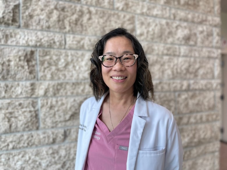 Dr. May Lynn Quan wears pink scrubs, a white lab coat and glasses. She's standing in front of a stone wall.