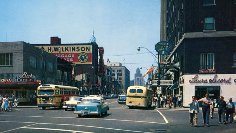 An old-timey photo shows people walking on sidewalks, cars and buses. 