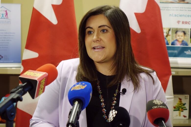 A woman with brown hair wearing a lavender suit jacket stands in front of 4 microphones.