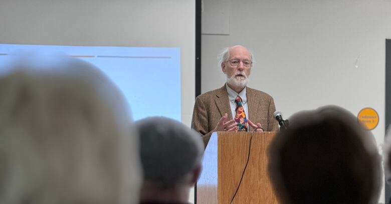 a gray haired bearded man wearing a suit and tie speaks from behind a podium to a crowd.