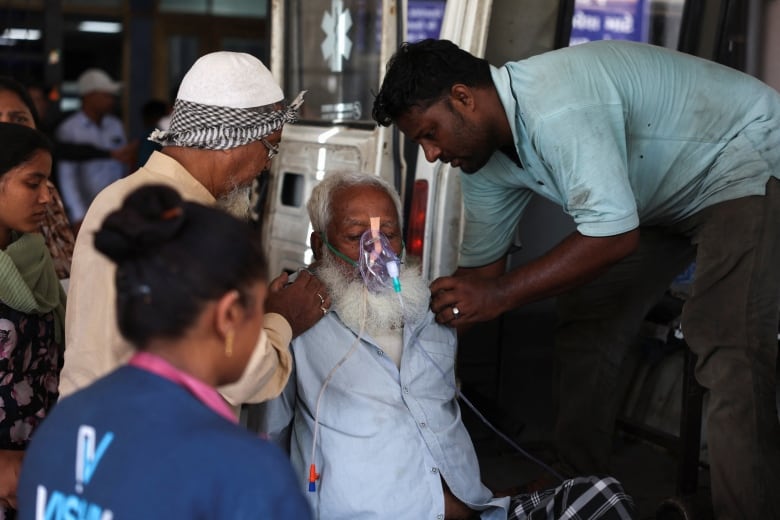 An elderly man with a white beard and an oxygen mask is helped by people surrounding him.